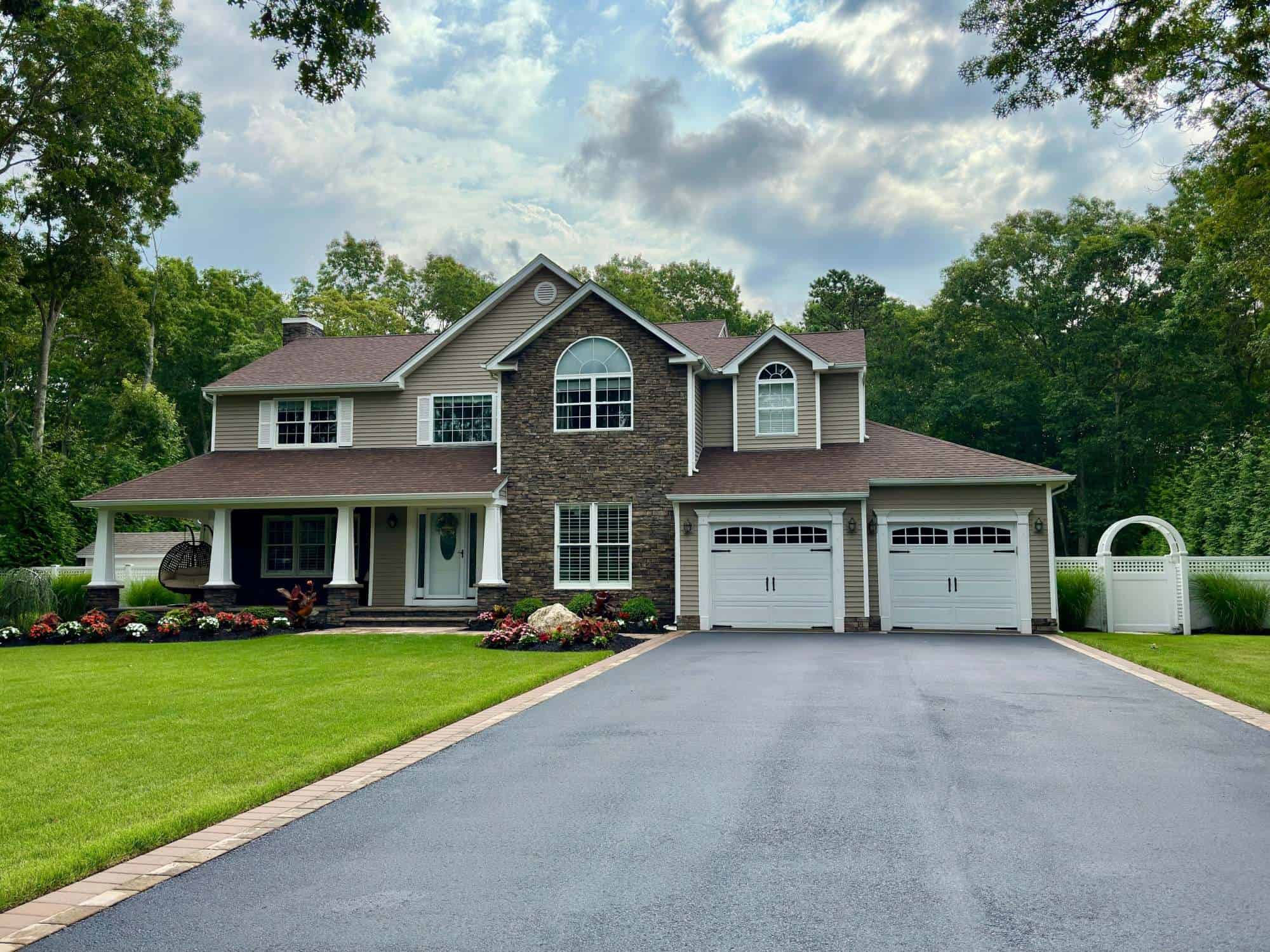 A two-story suburban home with brown brick and siding exterior, three white garage doors, and a lush green lawn. There are flower beds along the driveway and dense trees in the background under a cloudy sky.