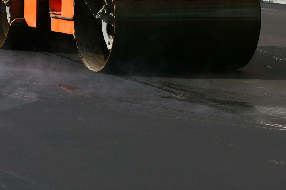 A steamroller compacts fresh asphalt on a road. The machine's large drum is partly visible, pressing the smooth, dark surface beneath it. Steam rises slightly, indicating the asphalt's new and hot condition.