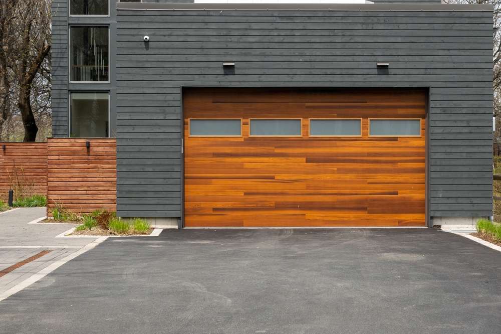 A modern house with a sleek, gray facade and a large wooden garage door featuring horizontal panels. The driveway is paved and lined with small plants. A wooden fence runs along the side of the house.