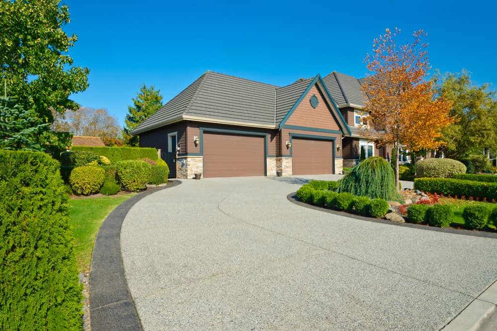 A modern suburban house with a large driveway, two brown garage doors, and a neatly manicured garden. The garden features shrubs, a small tree with autumn leaves, and a variety of greenery under a clear blue sky.