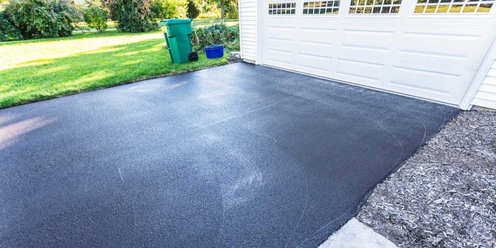 Freshly sealed driveway in front of a white garage with a large door. A green trash bin and blue container are nearby. The area is surrounded by bright green grass and trees in the background.