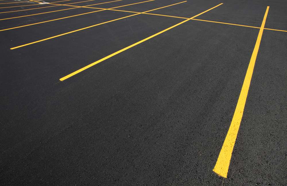 Empty parking lot with fresh black asphalt and bright yellow parking lines, creating a grid pattern. The surface appears smooth and well-maintained, with no vehicles present.