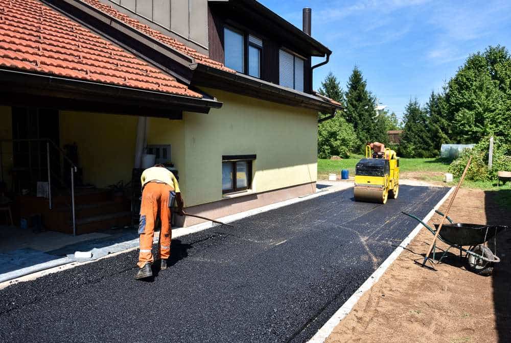 A worker in orange pants and a yellow vest is leveling fresh asphalt on a driveway beside a yellow house. A small yellow road roller is parked nearby. There are trees and a garden in the background under a clear blue sky.