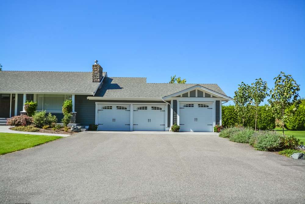 A suburban house with gray siding and a large driveway. It features a three-car garage with white doors and a small front garden with shrubs and trees. The roof is sloped and there are clear blue skies above.