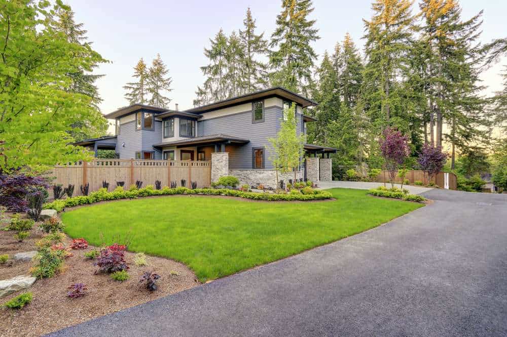 Modern two-story gray house with a stone facade, surrounded by a lush green lawn and landscaped garden. A paved driveway curves toward the entrance. Tall trees line the property, offering a serene, natural backdrop under a clear sky.