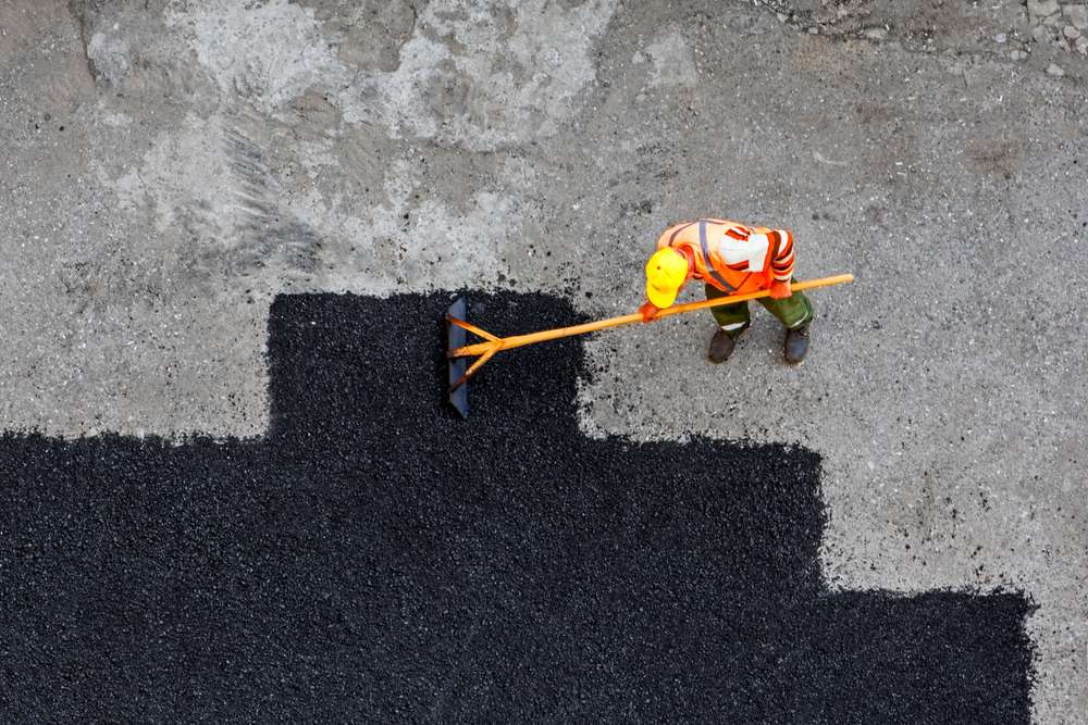 A worker wearing a yellow hard hat and orange safety vest spreads black asphalt with a large rake on a grayish surface. The contrasting colors emphasize the new section against the old pavement. Top-down view of the scene.