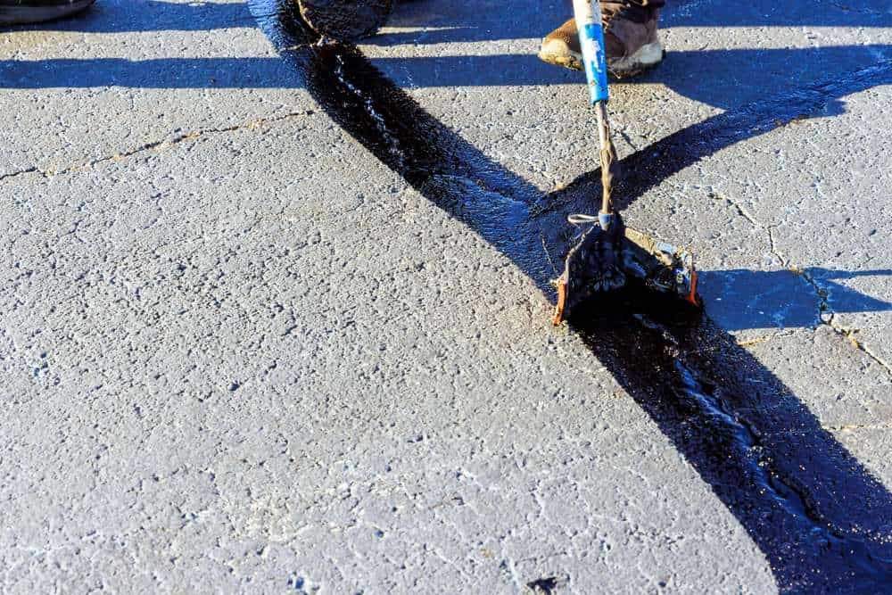 A person is applying black sealant to cracks in a gray asphalt surface using a long-handled tool. The textured road surface and shadows are visible in the bright sunlight.