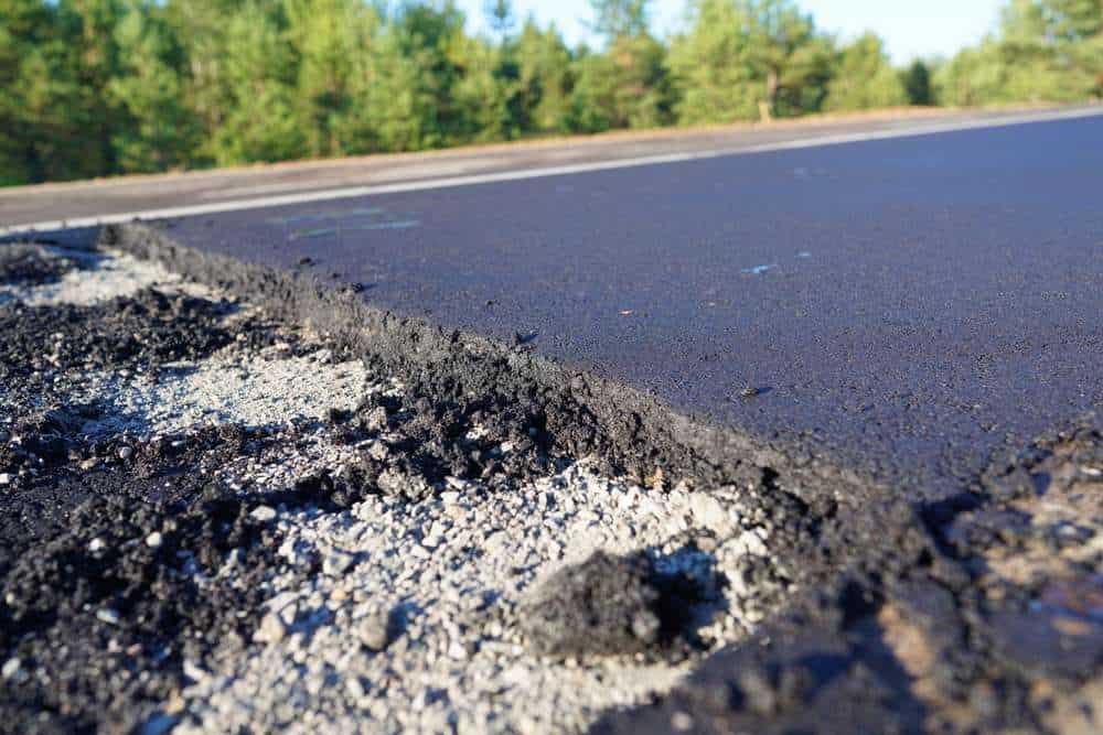 Close-up of a newly laid asphalt road showing a rough edge where it meets gravel. The background features green trees under a clear blue sky.