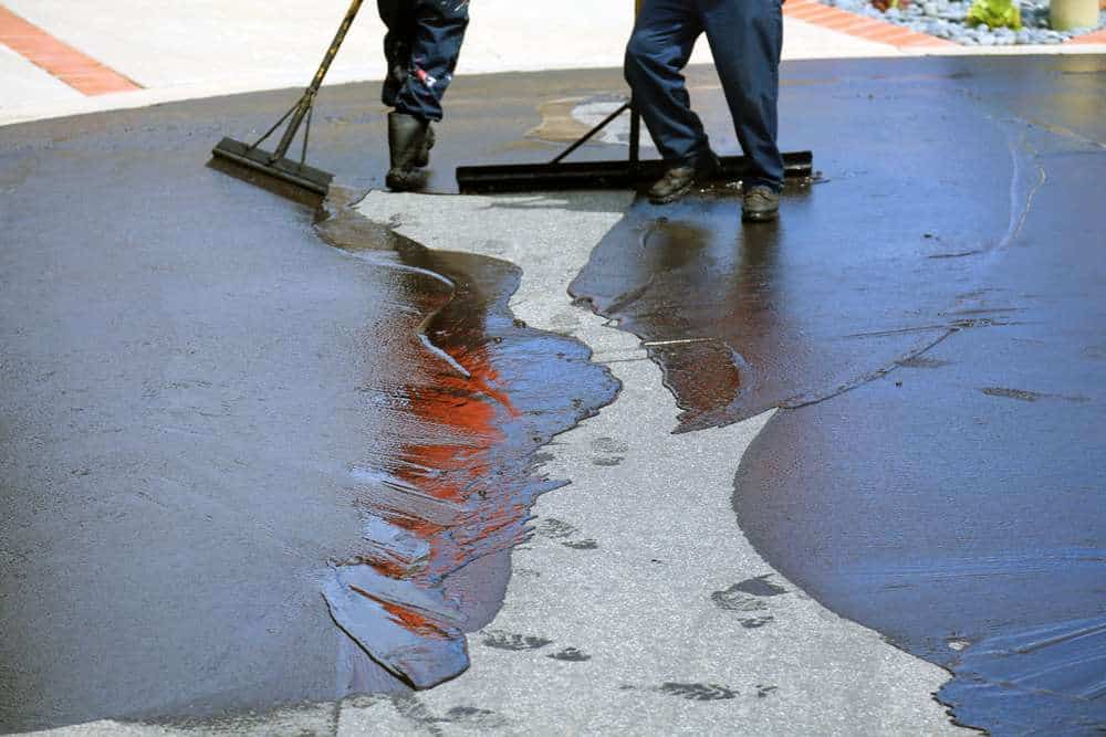 Two workers smooth fresh asphalt with large squeegees on a driveway. The asphalt is dark and shiny against the lighter background. One worker's boots leave footprints in the wet surface. The scene is outdoors, with some colored bricks visible.