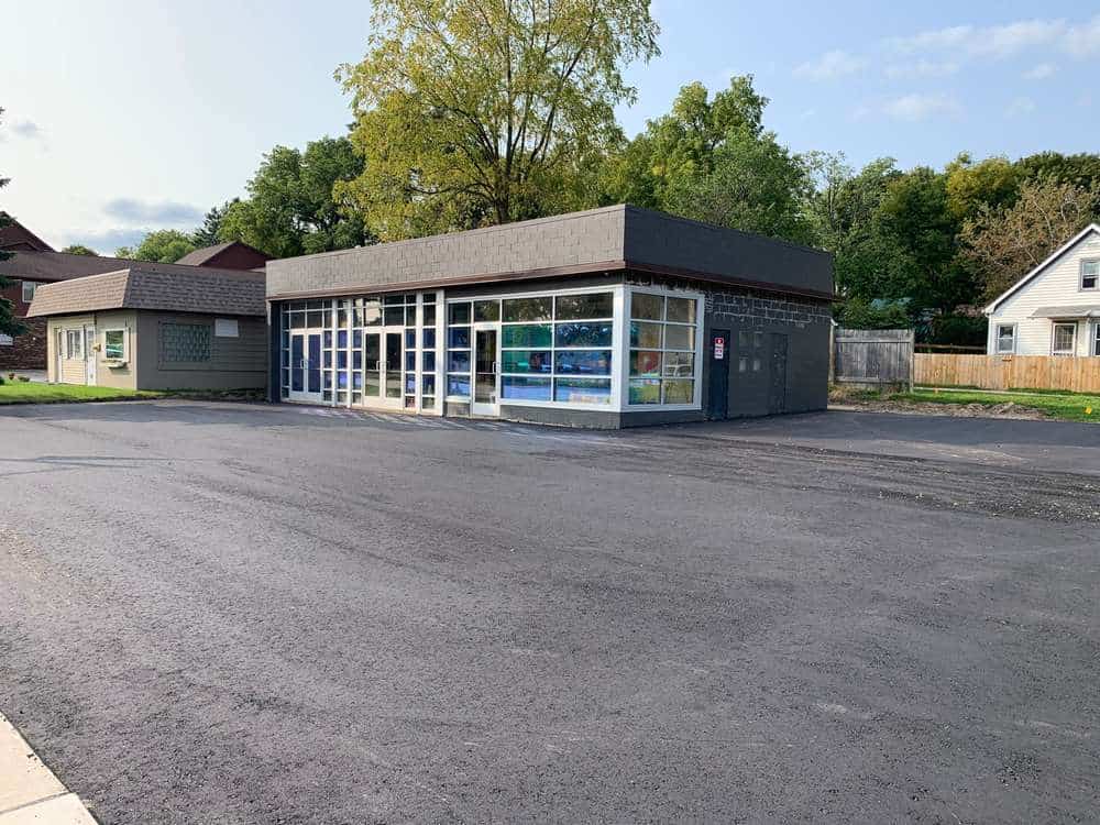 Small, modern commercial building with large windows and flat roof, set in an empty parking lot. A tree stands behind it, with more greenery and houses in the background under a partly cloudy sky.
