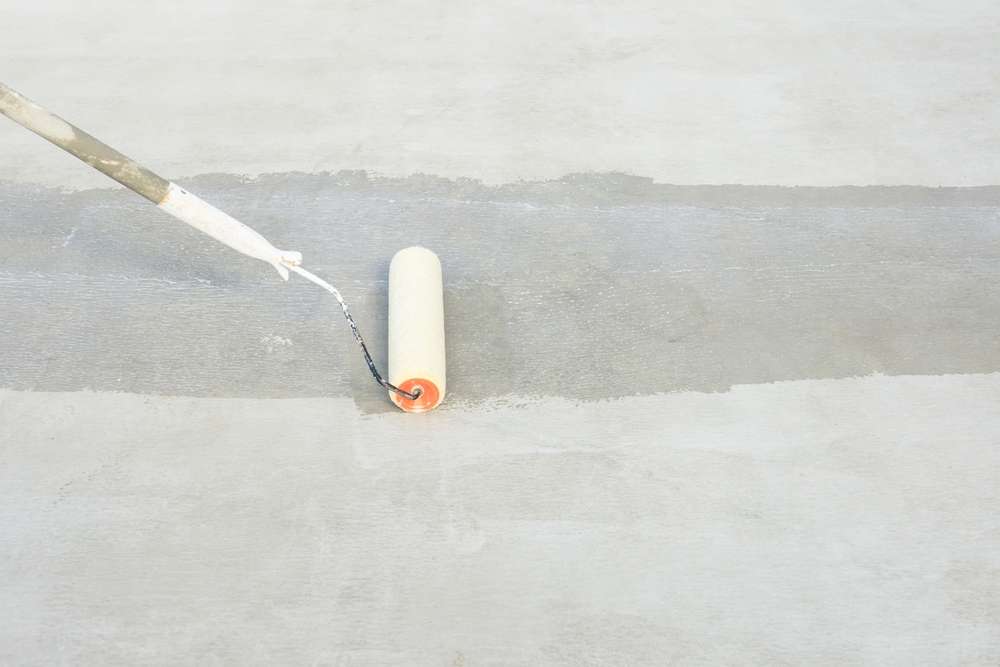 A paint roller on a long handle is rolling a light gray paint onto a smooth, light-colored surface, creating a neat painted strip in contrast to the surrounding area.