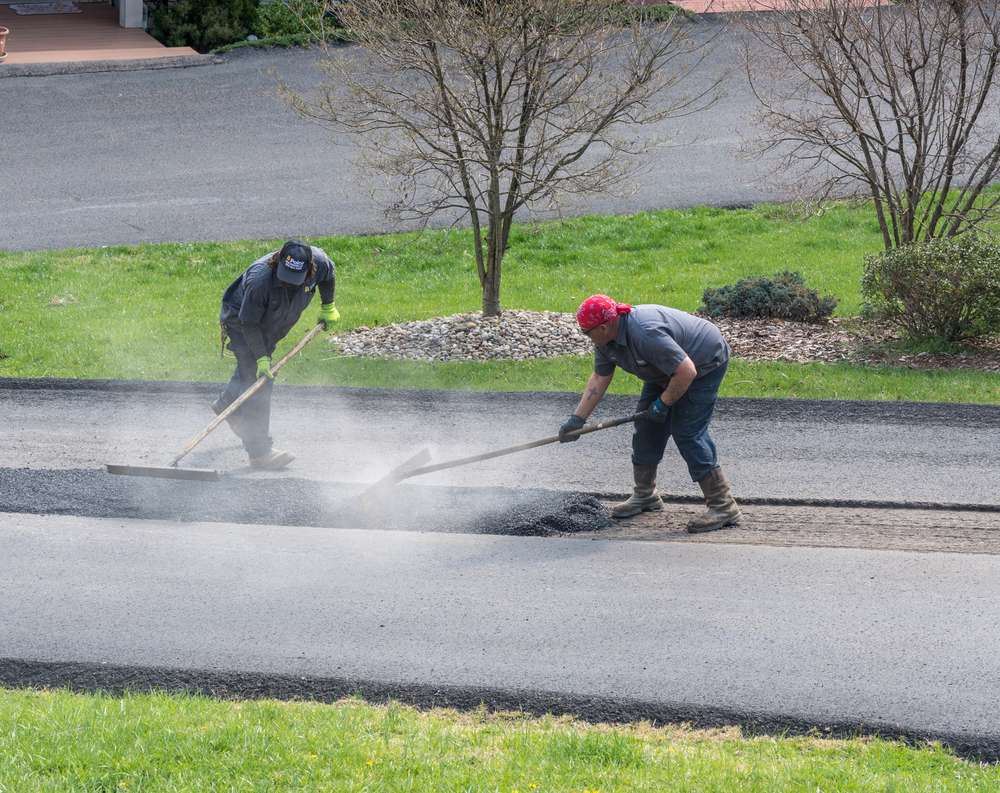 Two workers in protective gear paving a road with asphalt. One spreads the asphalt with a rake while the other smooths it with a tool. Dust rises around them on the freshly paved surface. Trees and grass are visible in the background.