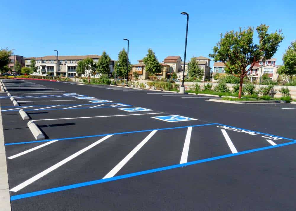 An empty parking lot with blue and white lines marking parking spaces, including accessible spaces near the front. Distant buildings and trees line the background under a clear blue sky.