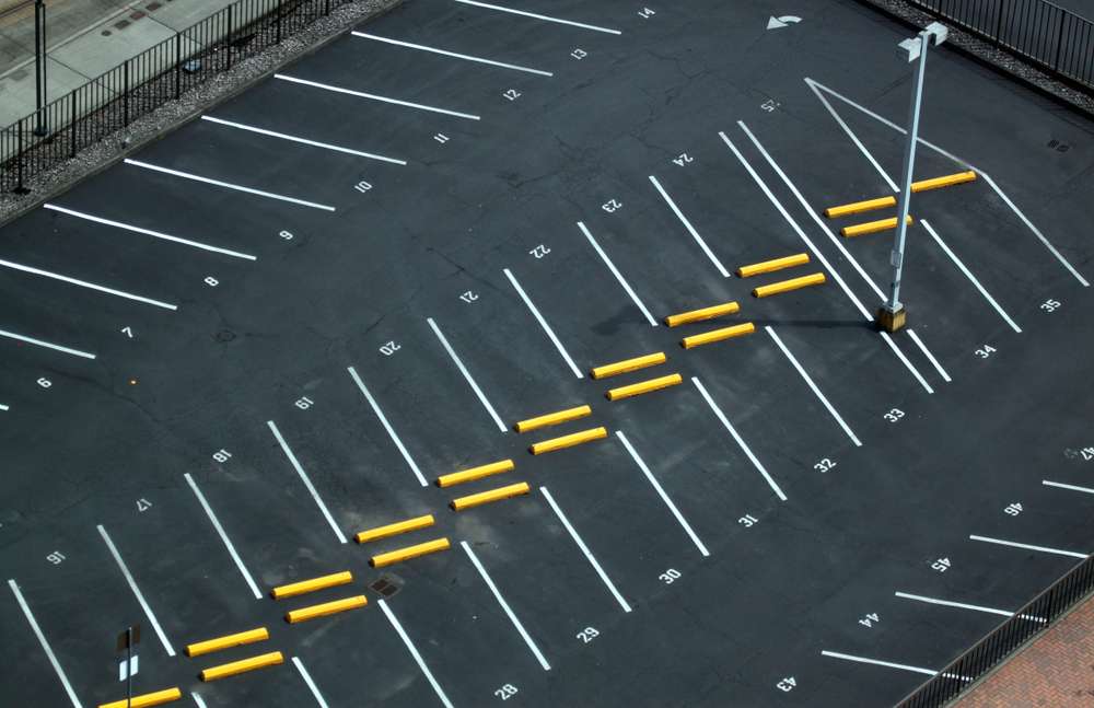 Aerial view of an empty parking lot with numbered spaces. Yellow barriers are evenly placed across the lot, and a tall light pole stands in the center. White lines are clearly marked for each parking spot.