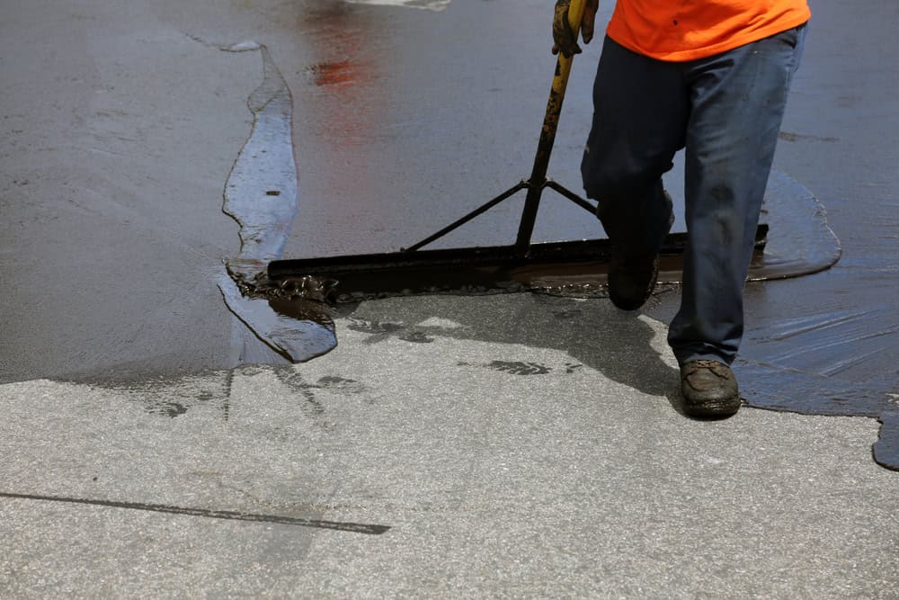 A person wearing an orange shirt and blue pants uses a tool to spread black asphalt during a roadwork project. Footprints are visible on the surface.
