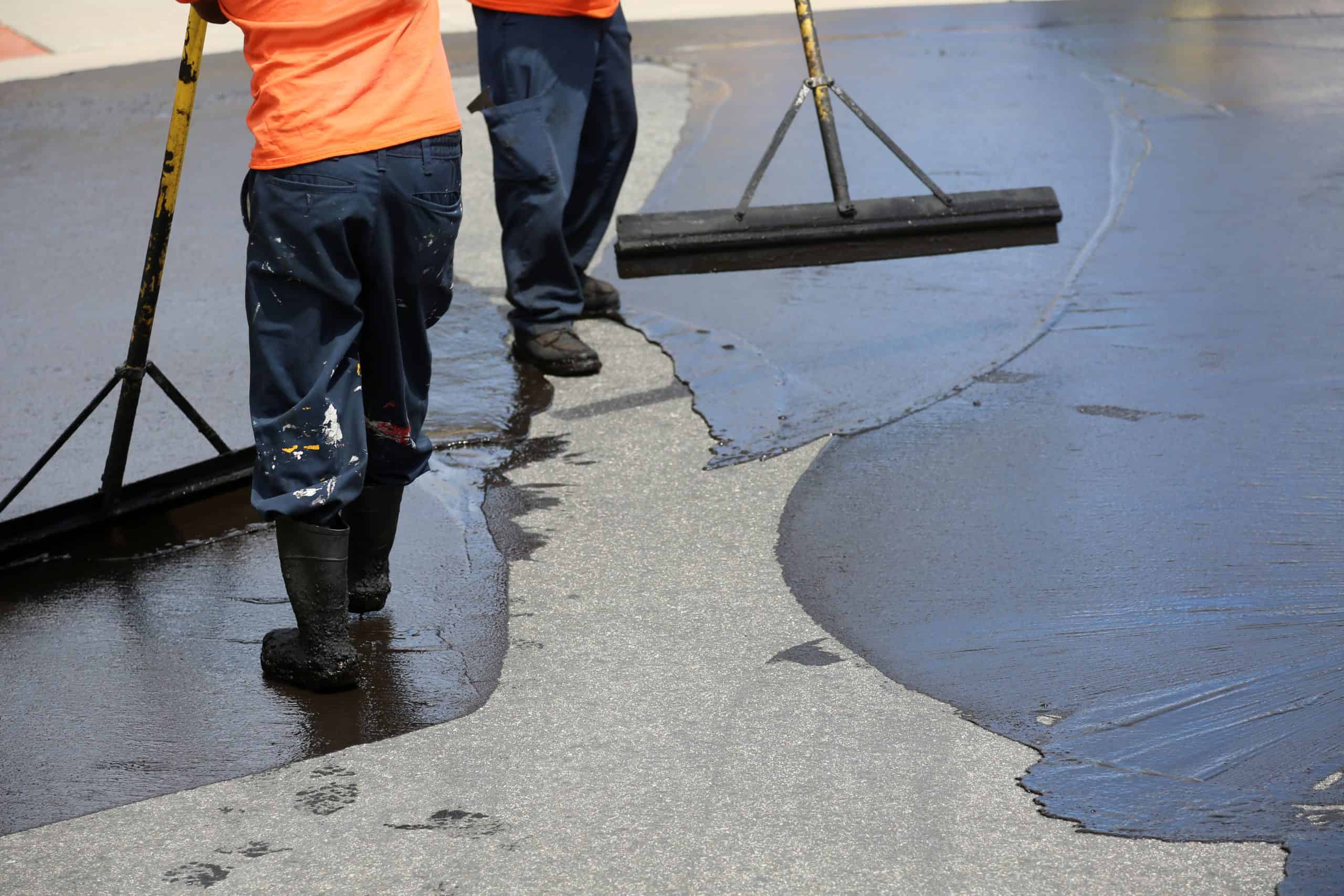 Two workers in orange shirts spread asphalt on a road using long-handled tools. They wear dark pants and black boots. The surface is partially covered with fresh asphalt, creating a smooth, dark finish as they apply the material.