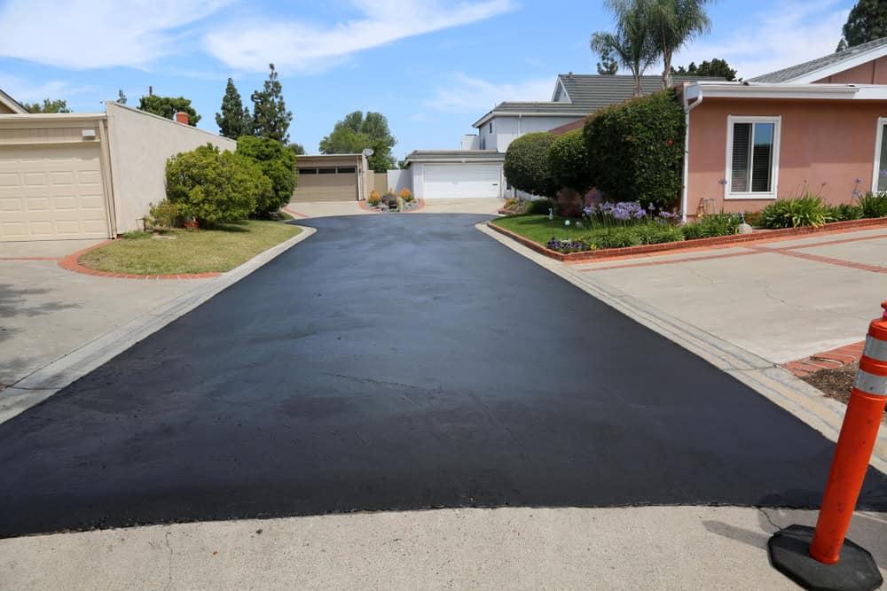 A freshly paved driveway leading to two garages, surrounded by manicured gardens and houses. A traffic cone is placed on the right side near the edge of the pavement. Blue sky and trees are visible in the background.