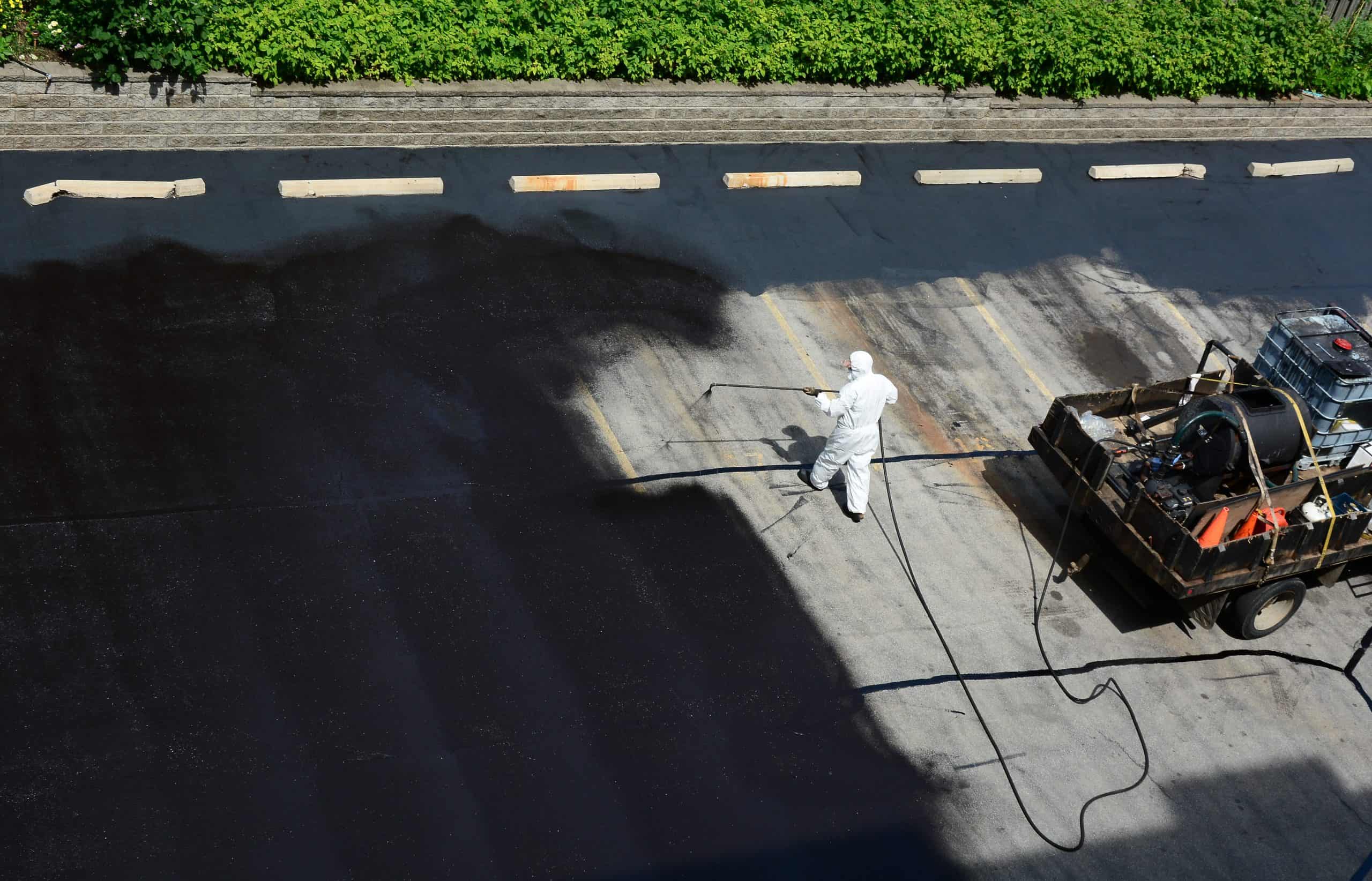 A worker in white protective clothing applies a dark sealant to a road surface using a spray apparatus. The truck carrying the equipment is nearby, and the treated portion contrasts with the untreated area. Green shrubs line the background.