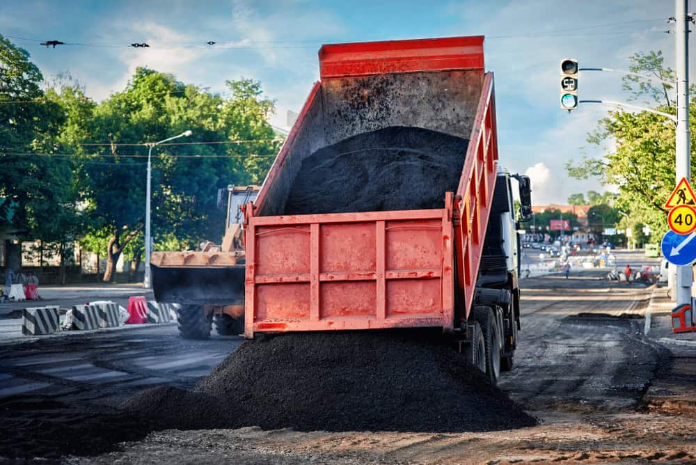 A red dump truck unloads a pile of black asphalt onto a road under construction. Traffic signs and barriers are visible, with a sunny blue sky and green trees in the background.