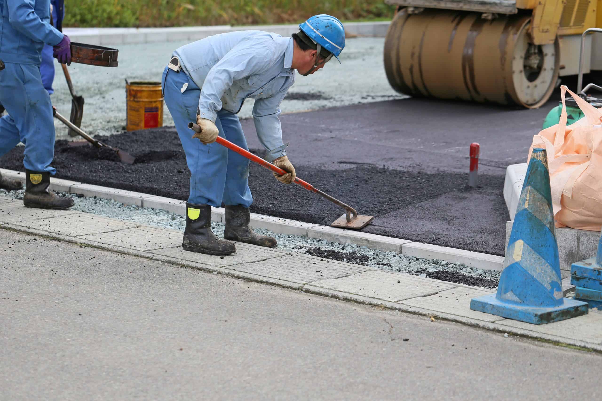 Construction workers in blue uniforms and helmets are spreading asphalt on a road with shovels. A roller machine is visible in the background. There are safety cones and a large bag on the side.