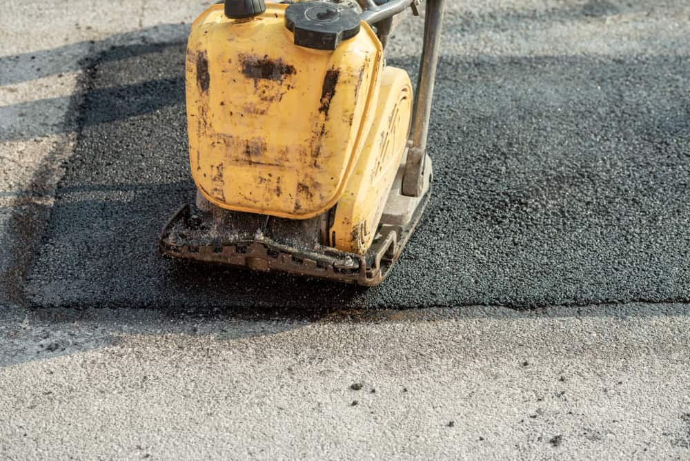 A yellow asphalt compactor machine pressing down freshly laid black asphalt on a road. The texture of the compressed asphalt contrasts with the surrounding area.