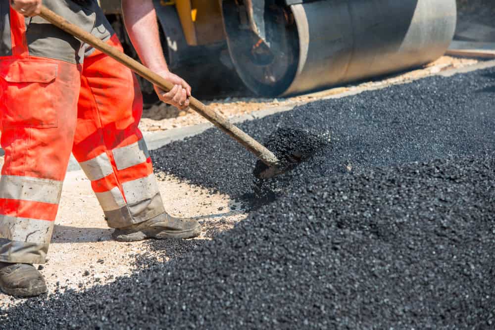 A worker in orange safety trousers shovels hot asphalt on a road construction site. A steamroller is visible in the background, helping to flatten and compact the surface. The scene is set during daylight.