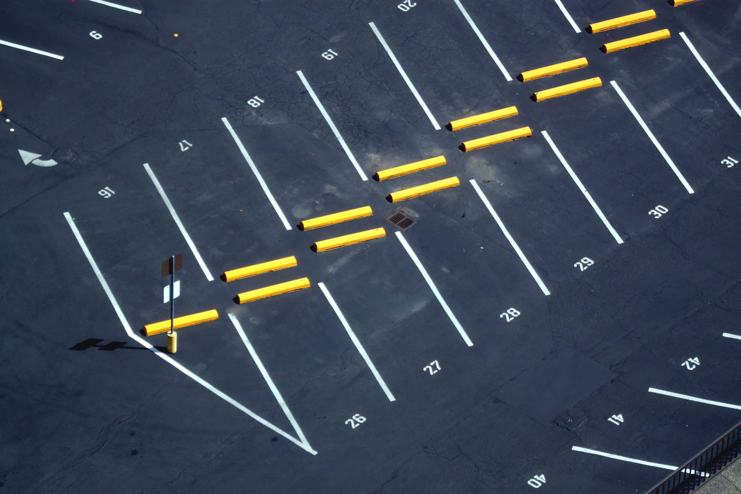 An empty parking lot with marked spaces, labeled numbers, and yellow wheel stops. The lot has visible cracks in the asphalt and arrows painted for direction.