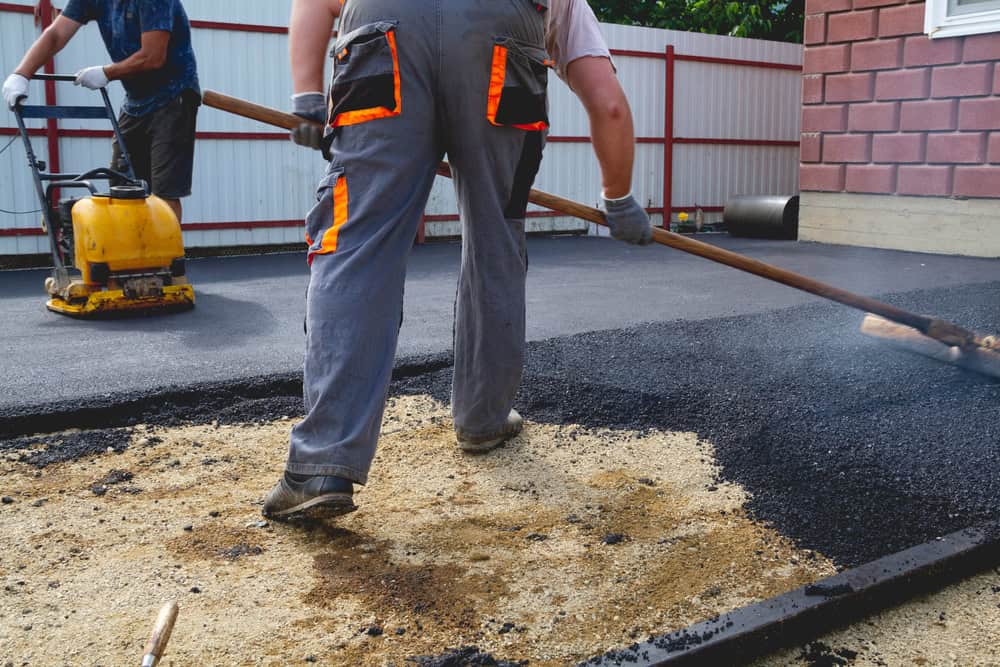 Two workers are paving a surface with asphalt. One is spreading the asphalt with a rake while another operates a yellow machinery. They are wearing protective gloves and work clothes. A metal and brick barrier is in the background.