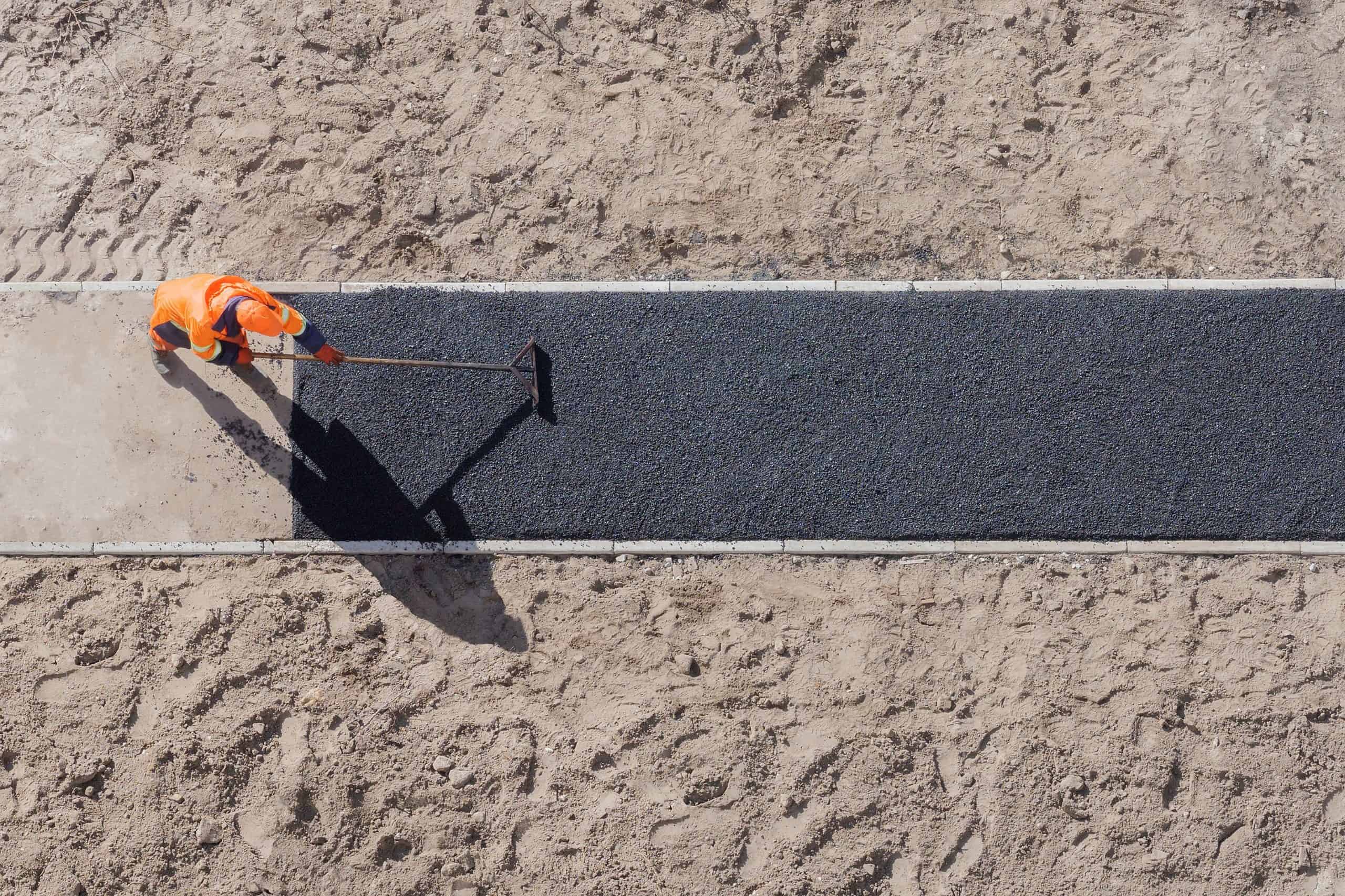 Aerial view of a construction worker in an orange safety outfit spreading asphalt on a path surrounded by sand. The worker is using a hand tool to level the surface, casting a shadow on the asphalt.
