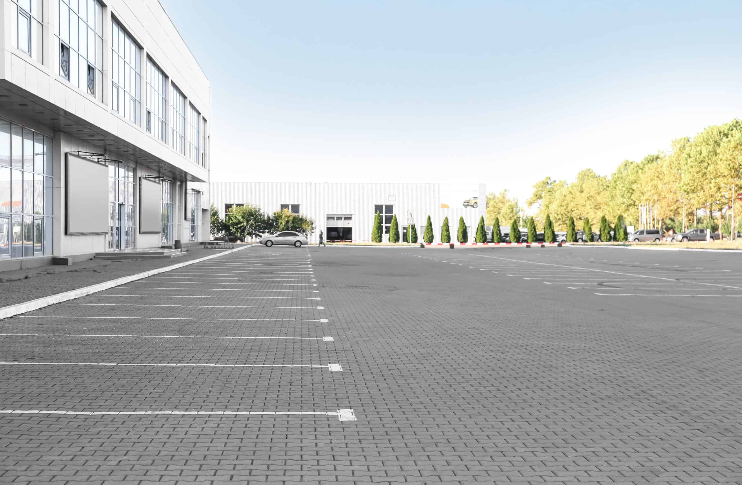 A large, empty parking lot with white-lined parking spaces in front of a modern building. Trees line the background with another building visible in the distance. The sky is clear and blue.