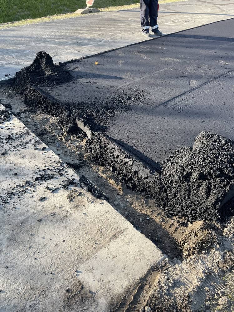 Fresh asphalt is being laid over a concrete surface, with visible sections of newly poured black asphalt. A person in dark pants and sneakers is partially visible near the top, standing on the new surface. Grass borders the area.
