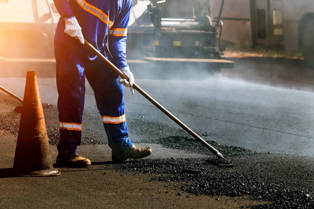 A worker in a blue uniform with reflective stripes spreads asphalt on a road using a long-handled tool. An orange traffic cone is nearby, and there's construction equipment in the background. Sunlight casts a warm glow over the scene.