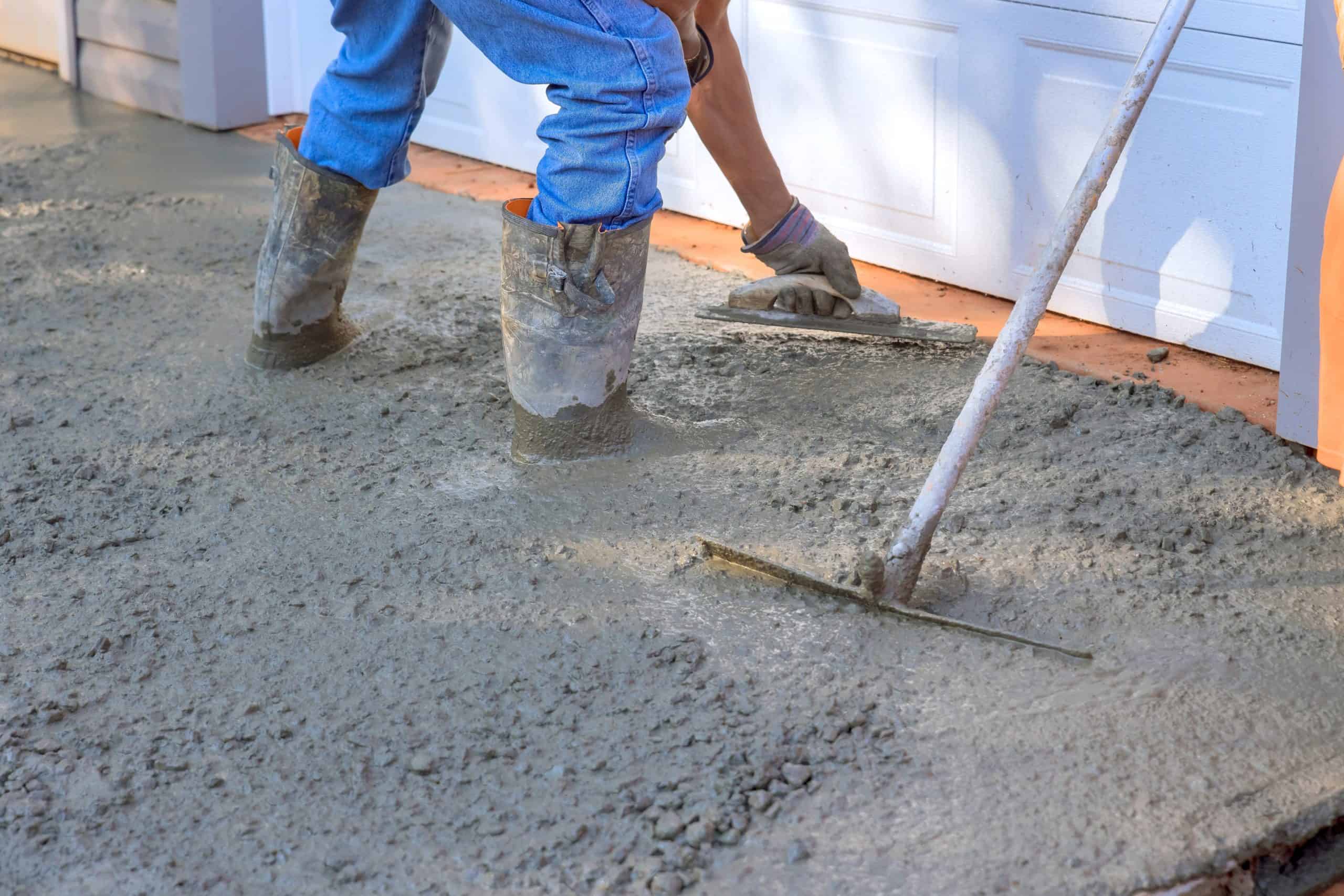 Two workers in boots and gloves smooth wet cement with a wooden trowel and long-handled tool, in front of a white building. The scene shows construction and paving work in progress.