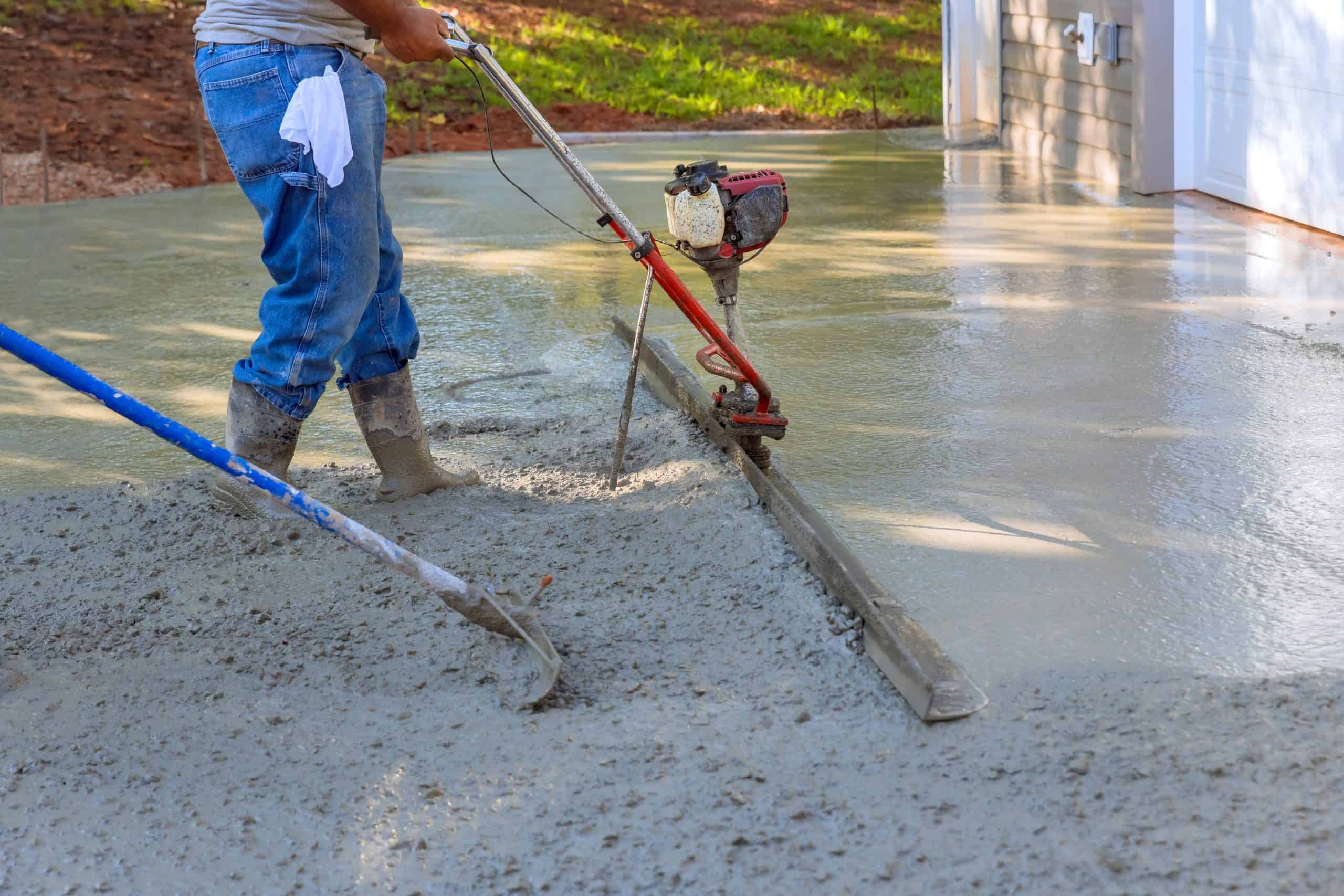 A person wearing jeans and boots is using a power trowel to smooth wet concrete in a driveway. Another tool with a blue handle is nearby. The concrete is fresh and still wet, surrounded by a grassy area.