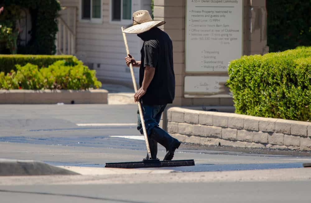 A person in a straw hat and dark clothing is applying sealant to a road with a large squeegee. They are working in a residential area, with green shrubs and a beige building in the background.