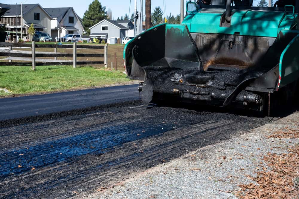 A green asphalt paving machine spreads fresh asphalt onto a residential road. The new asphalt contrasts with the surrounding gravel and older pavement. Houses and trees are visible in the background on a sunny day.