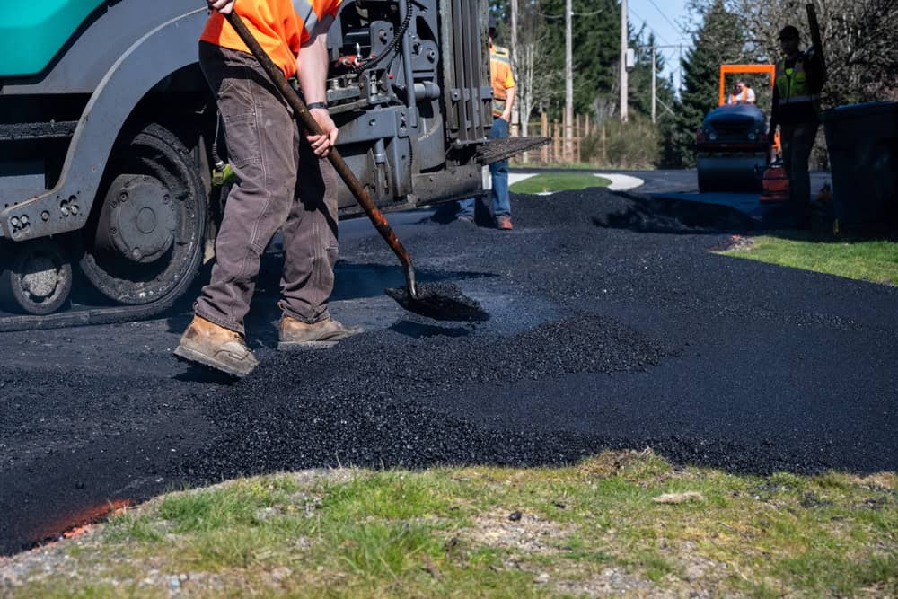 Workers in orange vests are paving a road. One uses a shovel to spread asphalt, while another operates machinery in the background. Freshly laid asphalt covers part of the road, contrasting with the green grass beside it.