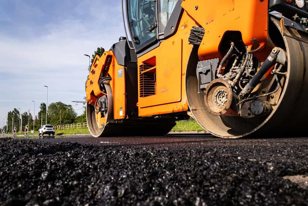 A close-up of an orange road roller compacting fresh asphalt on a road. The large, heavy wheels are pressing down on the black pavement. In the background, a white car is visible, and trees line the side of the road under a blue sky.