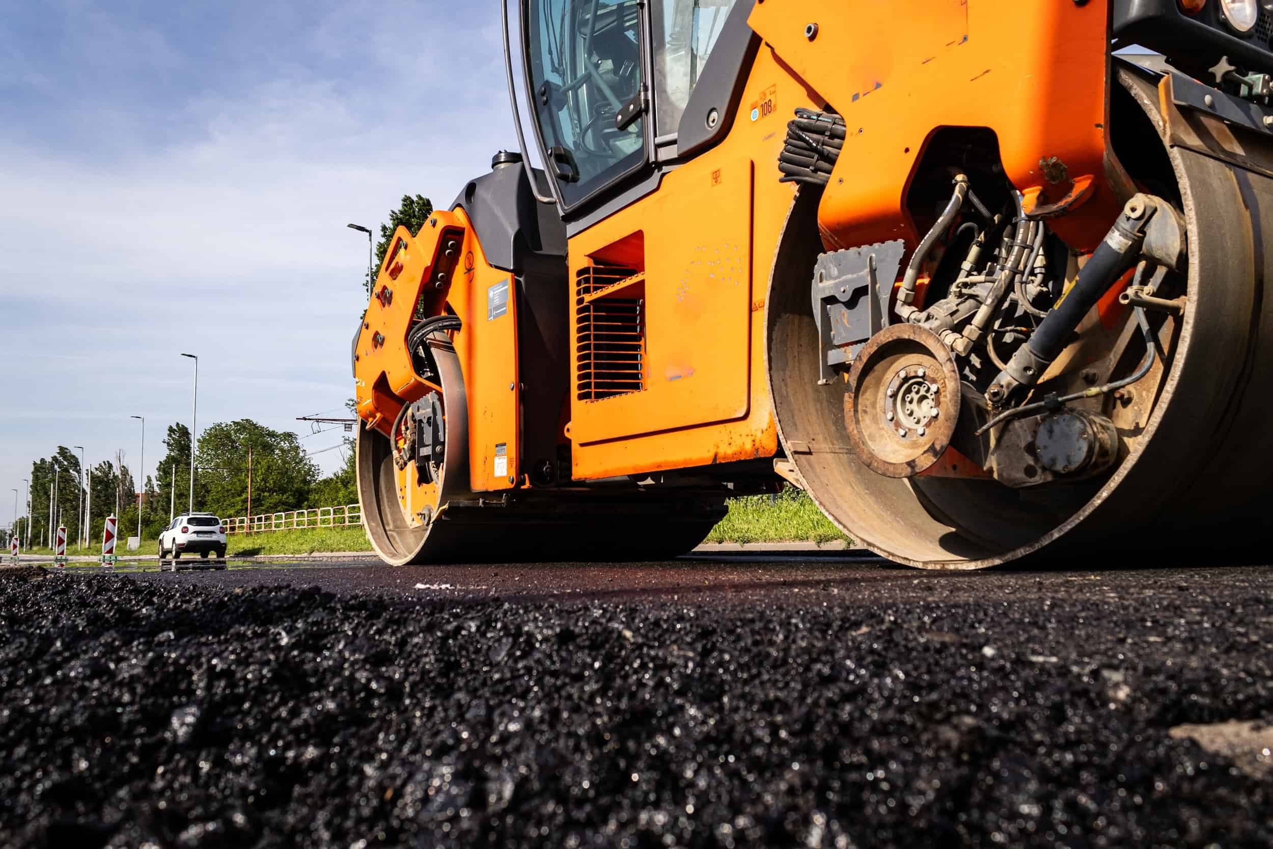 An orange road roller compacts fresh asphalt on a road under a clear sky. A white car and a few people are visible in the background. Green grass lines the side of the road.