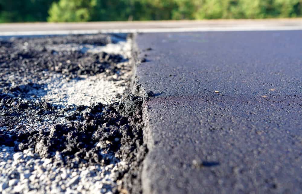 Close-up of a freshly laid asphalt road, showing the contrast between smooth black asphalt and rougher, lighter gravel on the side. The background contains blurred green trees under a clear sky.