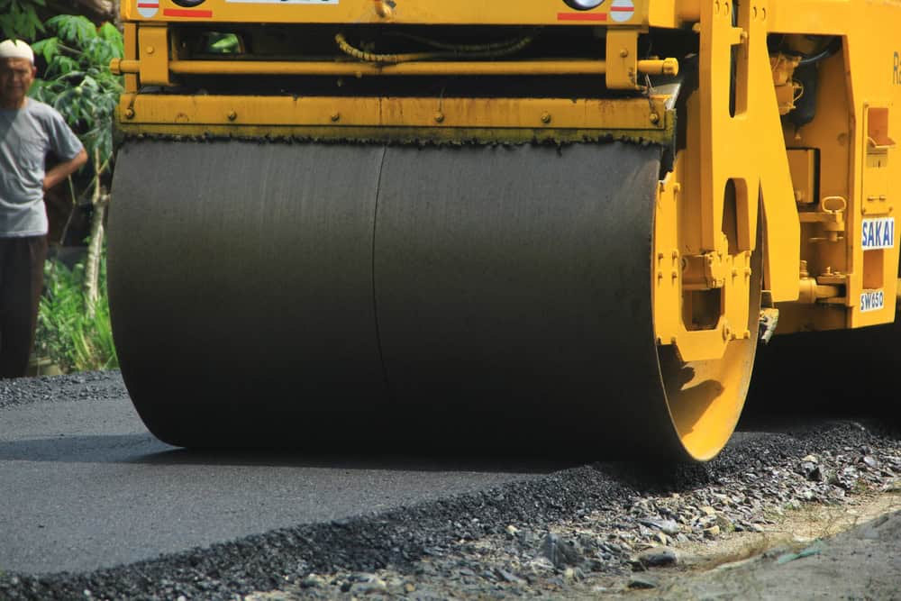A yellow steamroller compacts freshly laid asphalt on a road under construction. The large black roller is in motion. A person stands nearby watching the work.