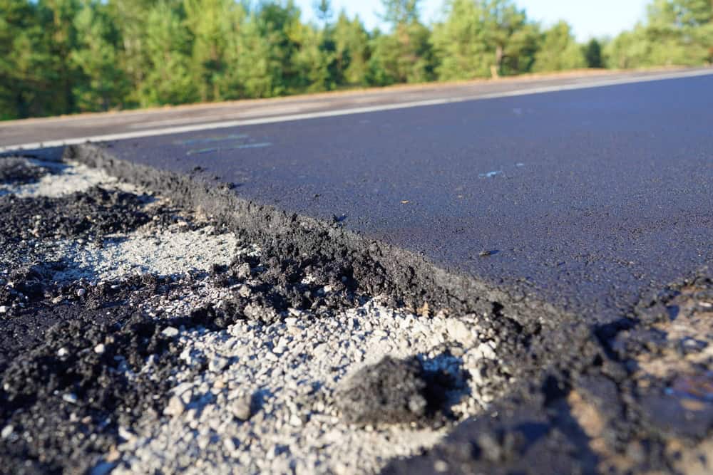 Close-up of a newly paved road with fresh asphalt. The edge of the asphalt meets a layer of crushed stone. Trees with green foliage are visible in the background under a clear blue sky.