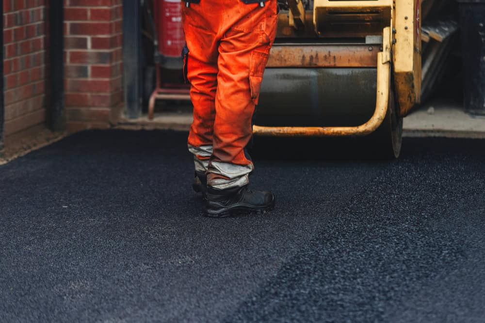 A construction worker in orange pants and black boots operates a yellow asphalt compactor on a freshly laid black surface next to a brick building wall.