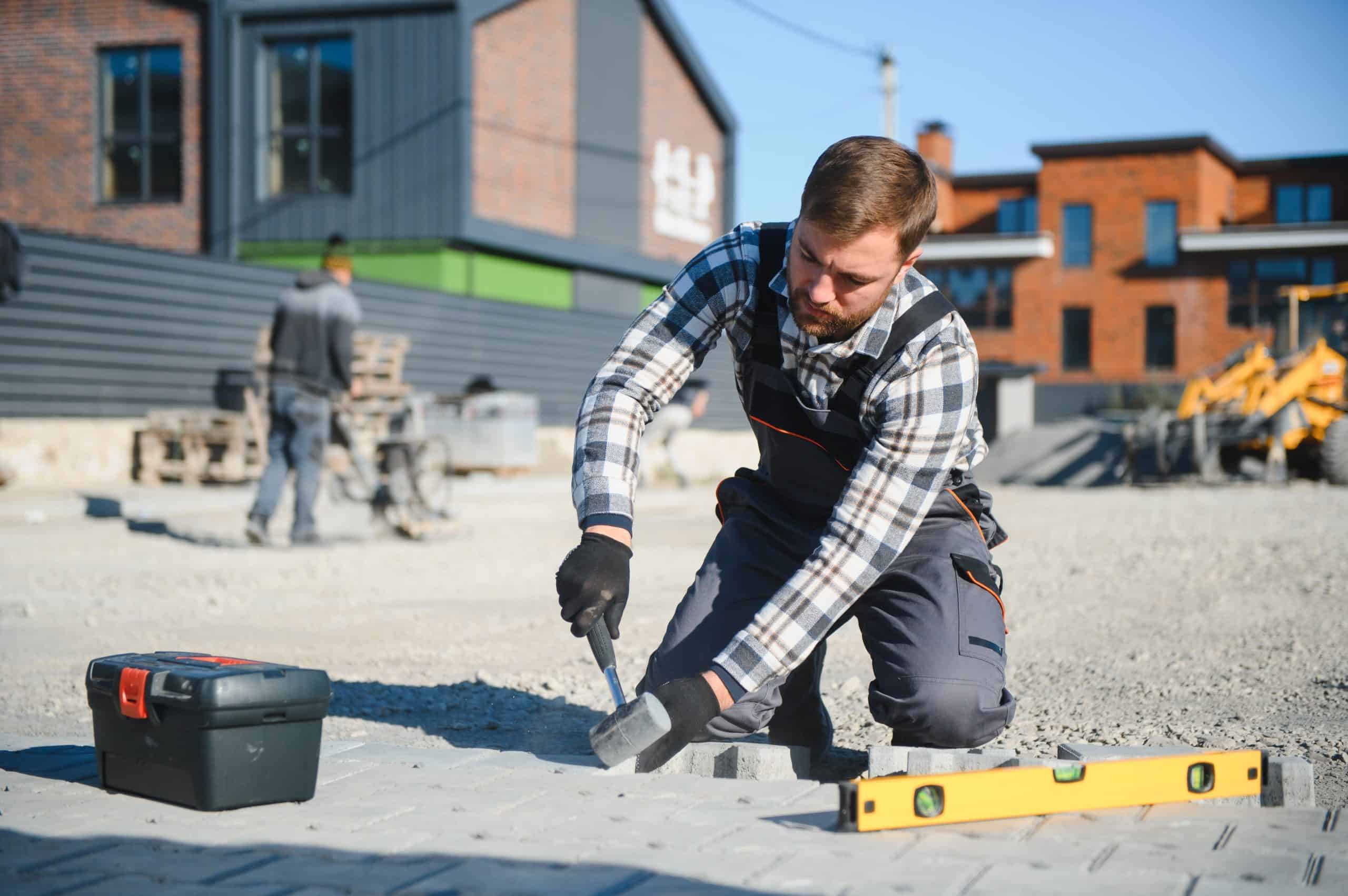 A construction worker in a plaid shirt and overalls kneels on a paved surface, using a hammer to adjust bricks. A spirit level and toolbox are nearby. In the background, there are brick buildings and construction equipment.