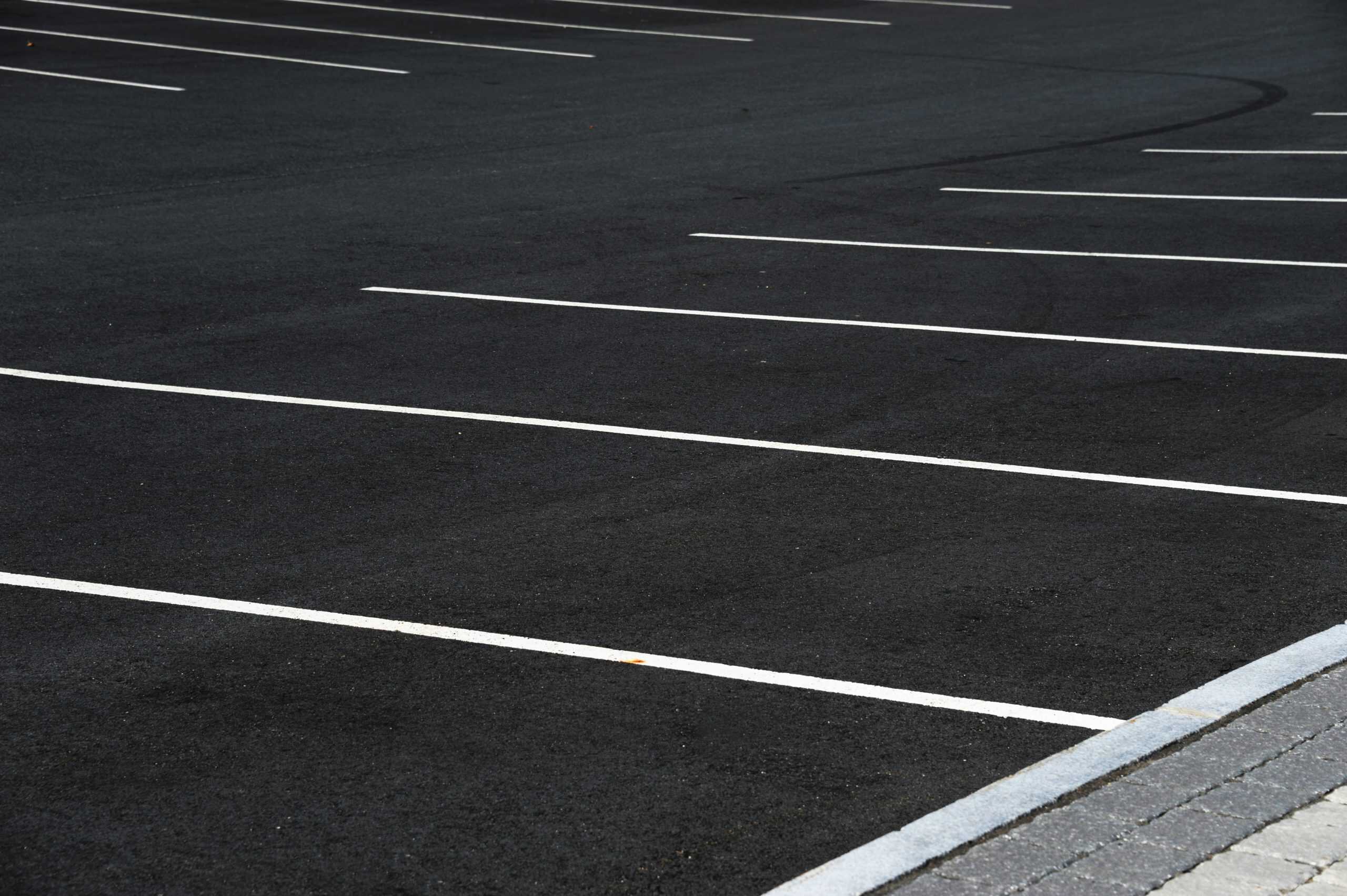 Empty asphalt parking lot with white painted lines marking individual spaces. There are no vehicles present. The foreground includes a section of sidewalk with light-colored paving stones.