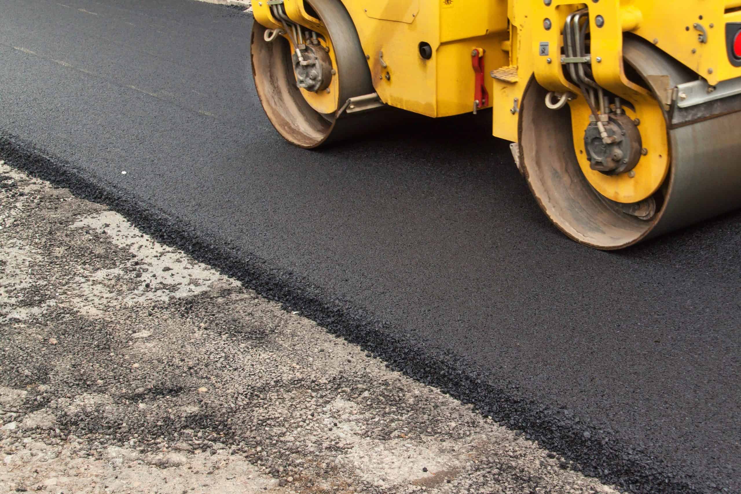 A yellow road roller compacts fresh black asphalt on a road, with the left side unfinished and rough. The roller's large, round drums press down on the asphalt, ensuring a smooth, level surface.