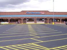 An empty parking lot with yellow lines in front of a brick grocery store, featuring a blue awning and a cloudy sky in the background&mdash;a fine example of expert parking lot paving by contractors from Hillsborough County, NH.