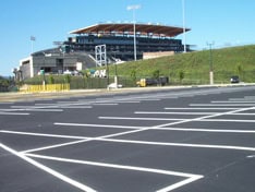 An empty, freshly painted parking lot with crisp white lines stretches into the foreground, showcasing the expertise of paving contractors in Cheshire County, NH. In the background, a large stadium with seating and a partial roof rises against a clear blue sky.