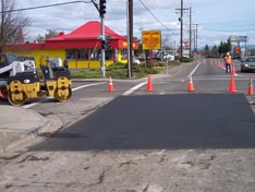 A freshly paved road section is cordoned off with orange traffic cones. A construction worker from a reputable paving contractor in Hillsborough County, NH stands nearby, and a small bulldozer is parked on the side. In the background, there is a yellow and red building and a traffic light.