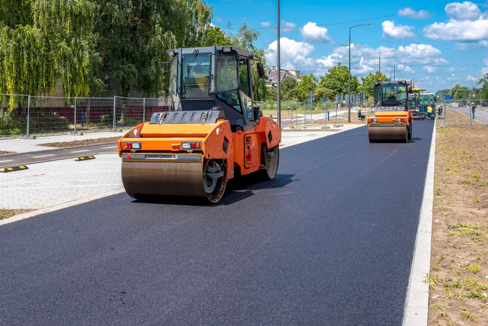 Two orange road rollers compact freshly laid asphalt on a street. The machines are working in a straight line under a sunny sky, and trees are visible in the background. A sidewalk with a fence runs parallel to the road on the left.